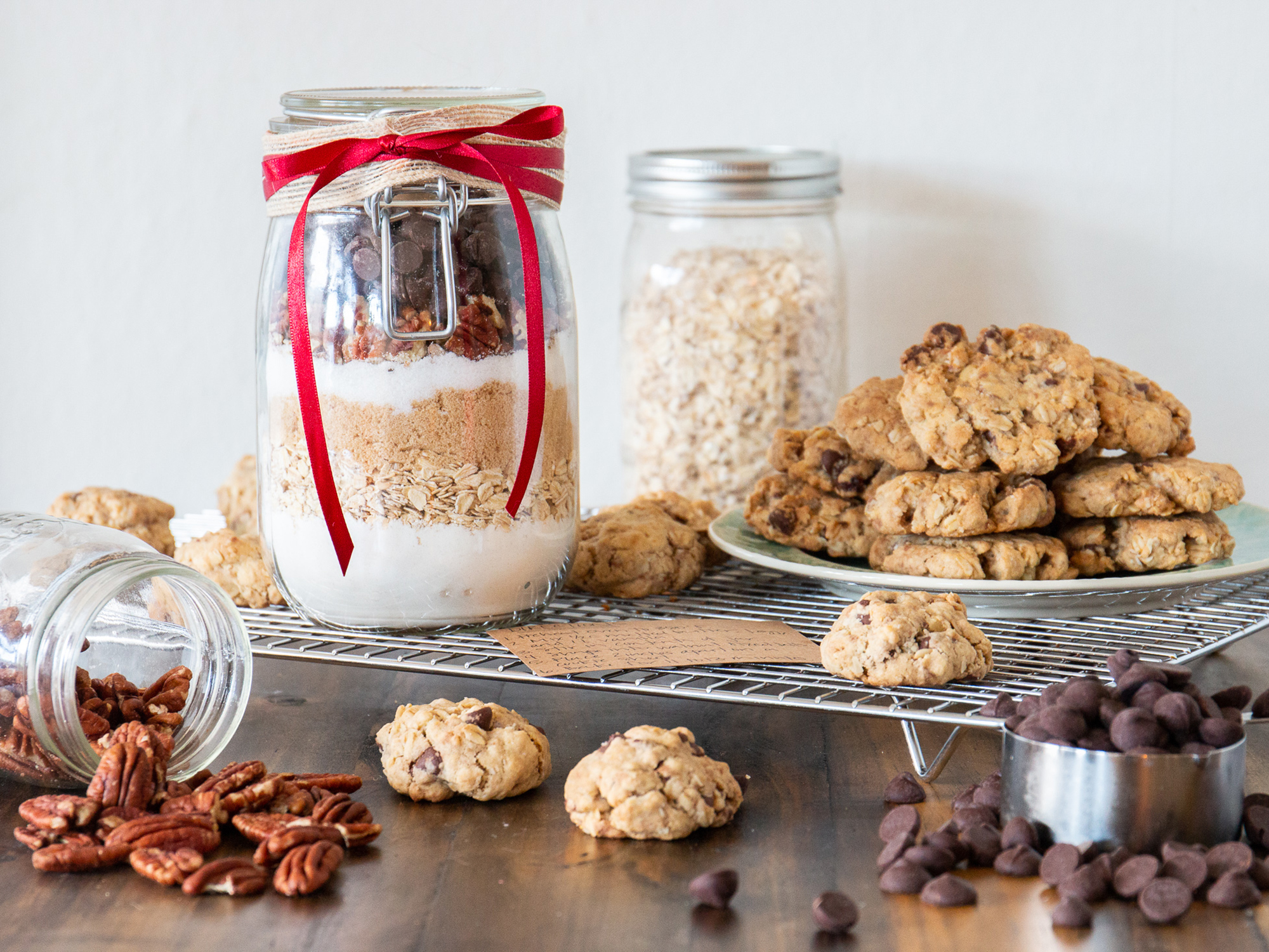 close up view of Cowboy Cookie Mix in a Jar with a red bow, next to cookies on a plate, nuts in a jar and chocolate chips in a measuring cup