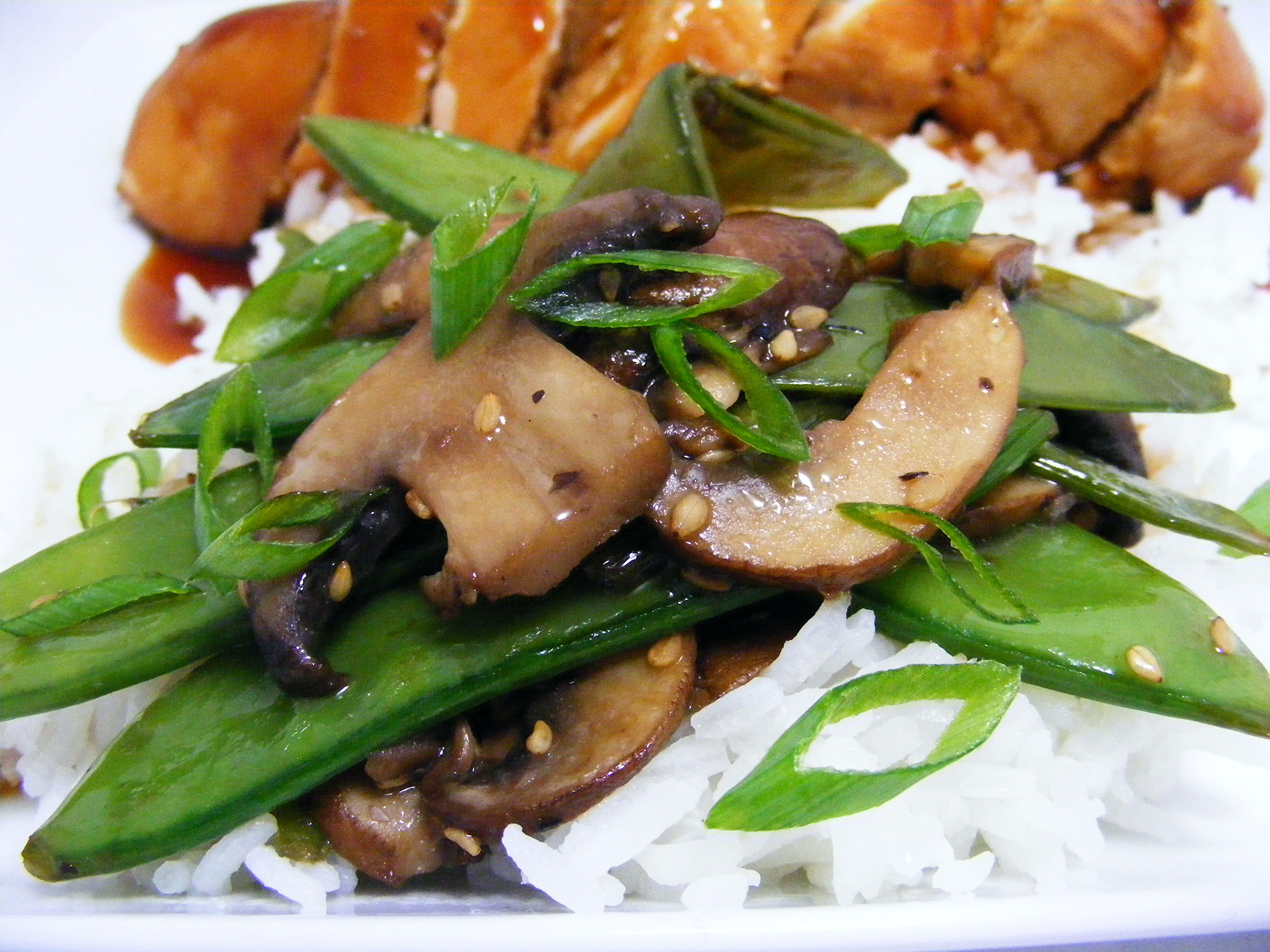 close up view of Stir Fried Snow Peas and Mushrooms garnished with green onions, over white rice, served with chicken on a white plate