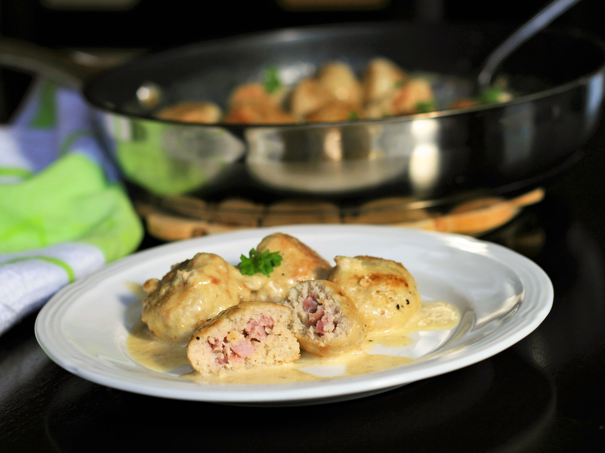 close up view of Keto Chicken Cordon Bleu Meatballs garnished with fresh herbs on a white plate, and Keto Chicken Cordon Bleu Meatballs in a pan in the background