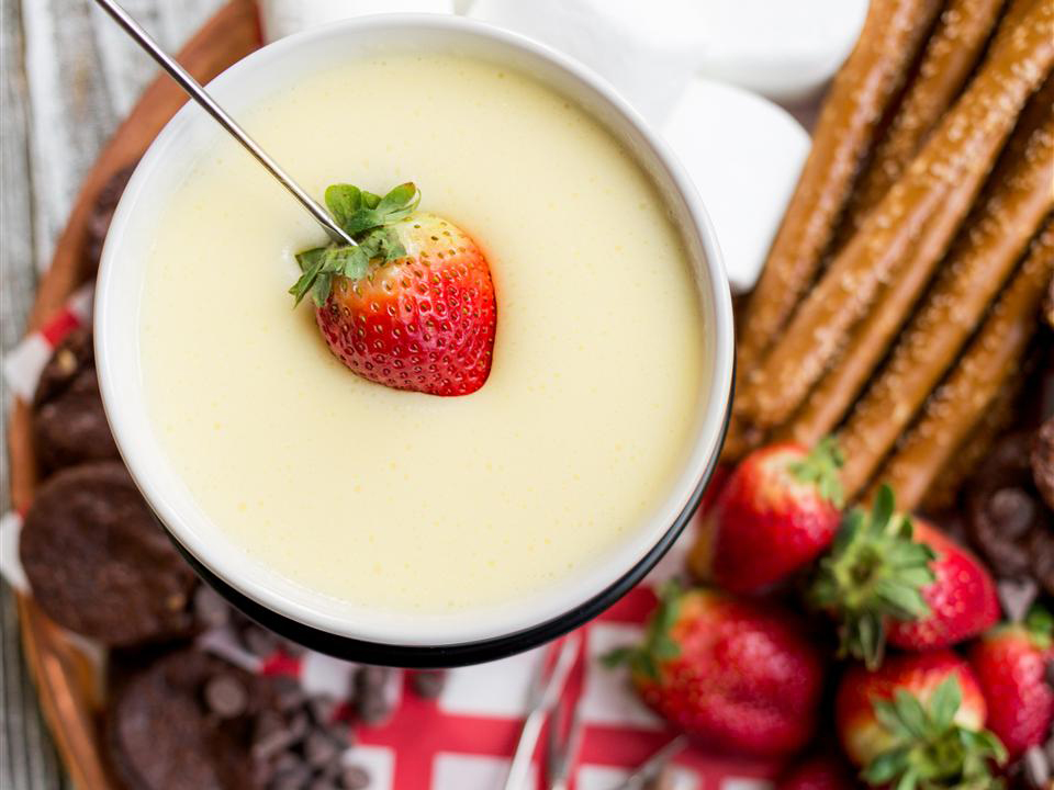 close up view of White Chocolate Fondue with a strawberry dipped inside, served with marshmallows, pretzel sticks, strawberries and chocolate