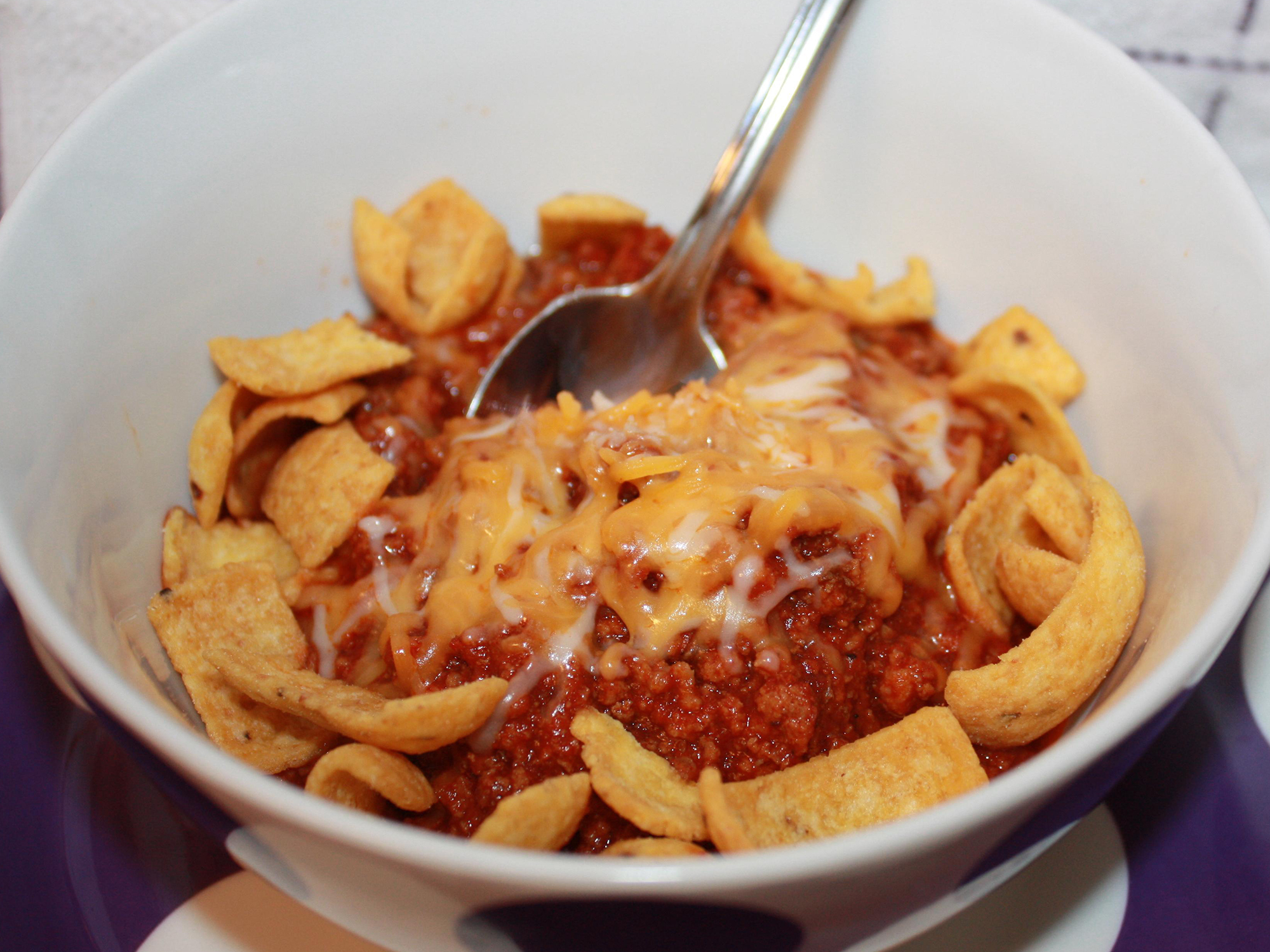 close up view of Skyline Chili garnished with cheese and corn chips in a white bowl with a spoon