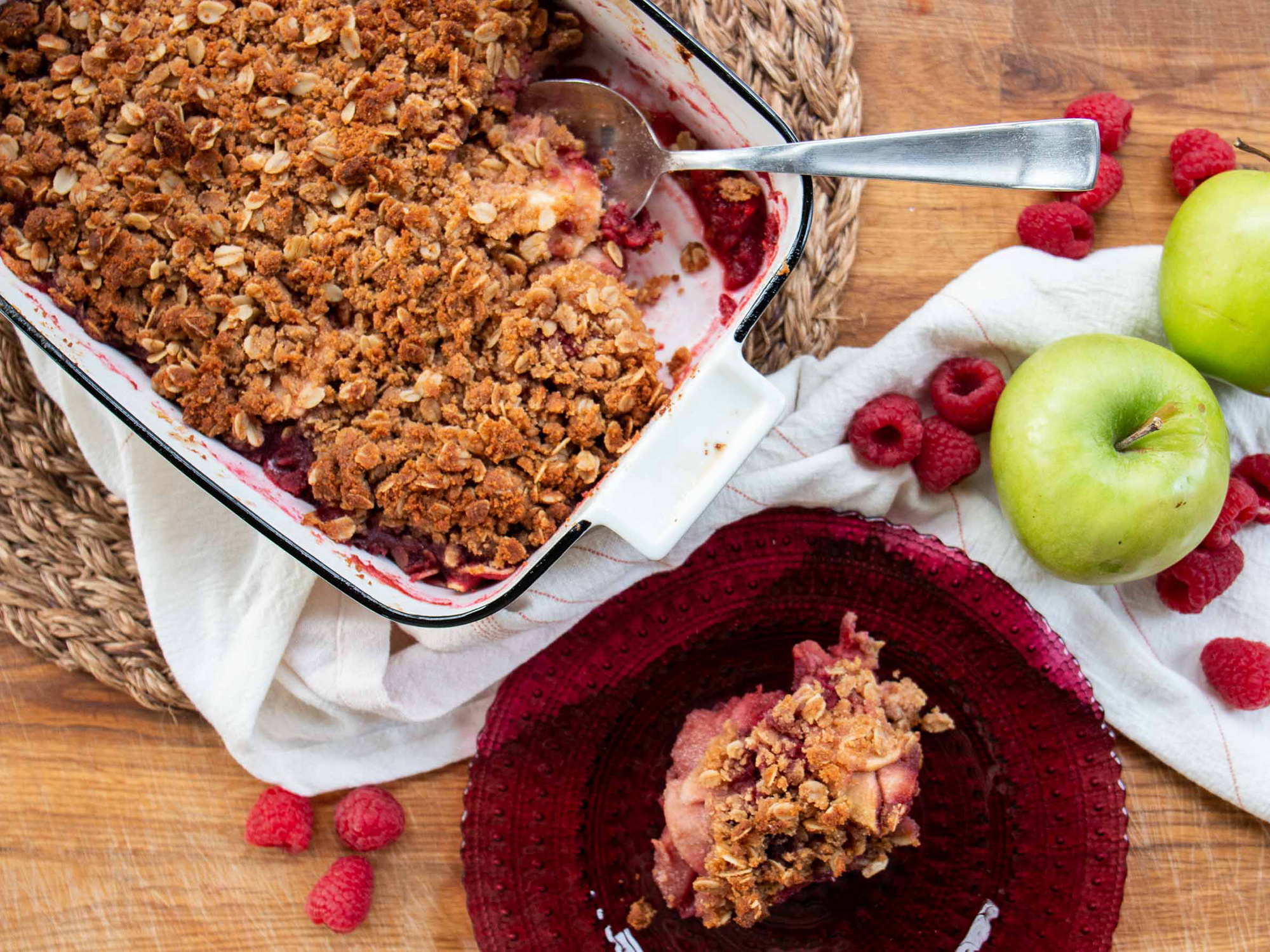 close up view of a scoop of Apple-Raspberry Crisp on a plate, and a Apple-Raspberry Crisp with a serving spoon in a baking dish, next to raspberries and apples