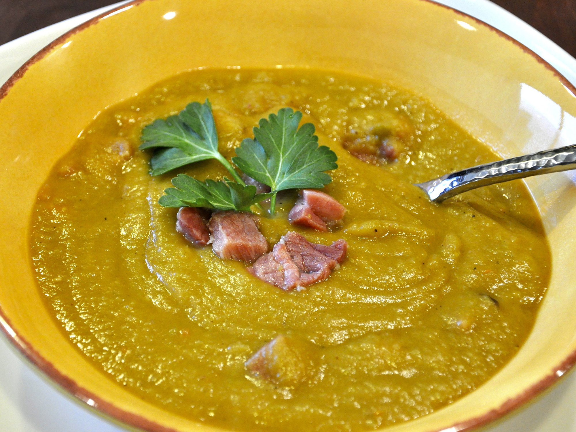 close up view of Split Pea Smoked Turkey Soup garnished with smoked turkey and fresh herbs in a yellow bowl, with a spoon