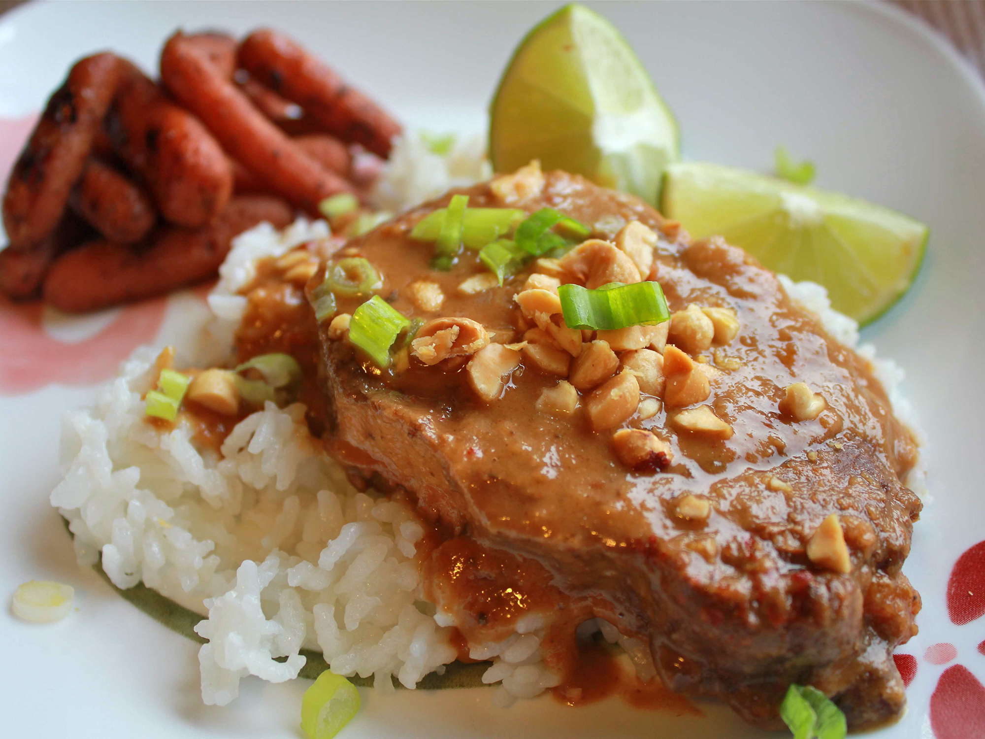 close up view of Thai Peanut Pork over white rice, garnished with peanuts and green onions, served with lime wedges and carrots on a plate