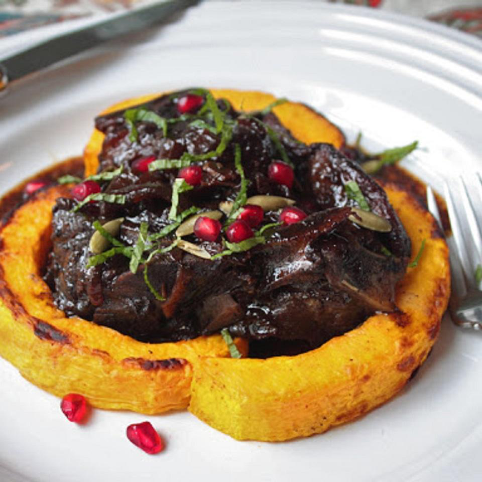close up view of Lamb Braised in Pomegranate served with baked pumpkin, garnished with fresh herbs, pumpkin seeds and pomegranate seeds on a white plate