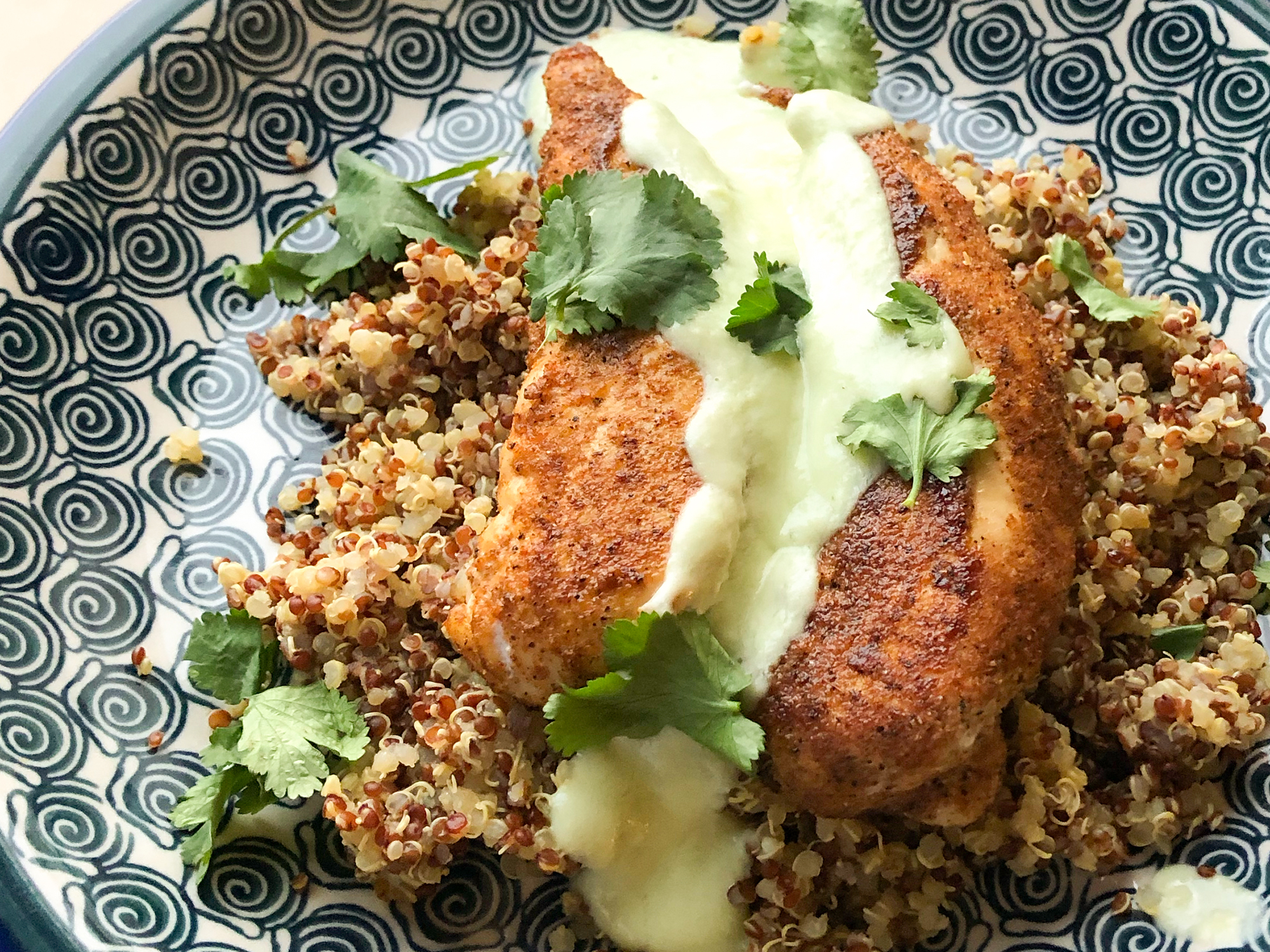 close up view of Blackened Chicken with Avocado Cream Sauce over quinoa, garnished with fresh herbs, on a plate