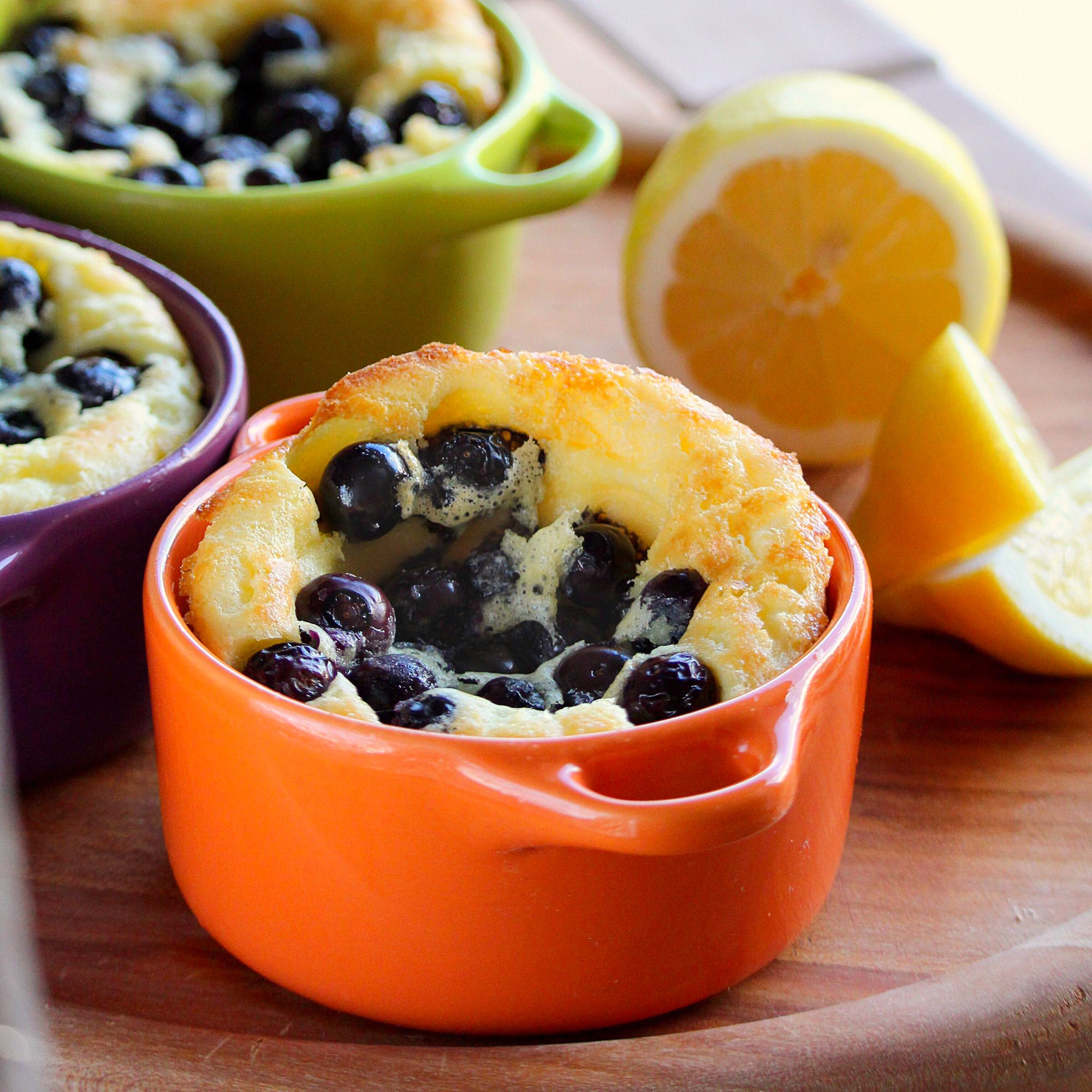 close up view of Blueberry Popovers in orange, purple and green ramekins, next to lemon wedges on a wooden board