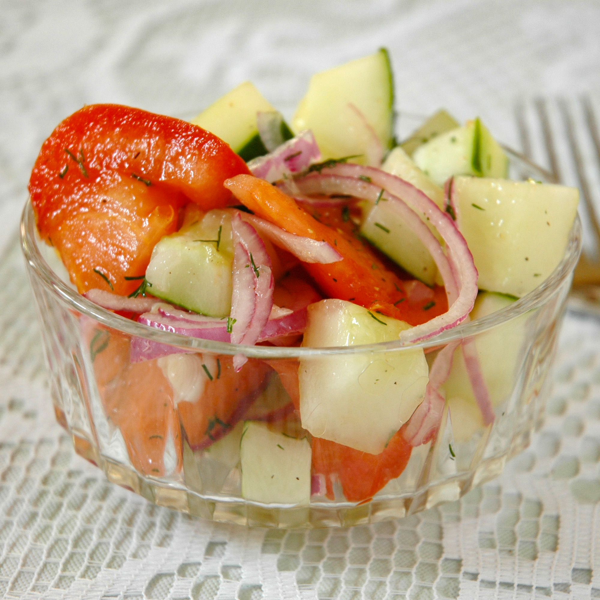 close up view of a Crispy Cucumbers and Tomatoes in Dill Dressing with red onions in a glass bowl, next to a fork