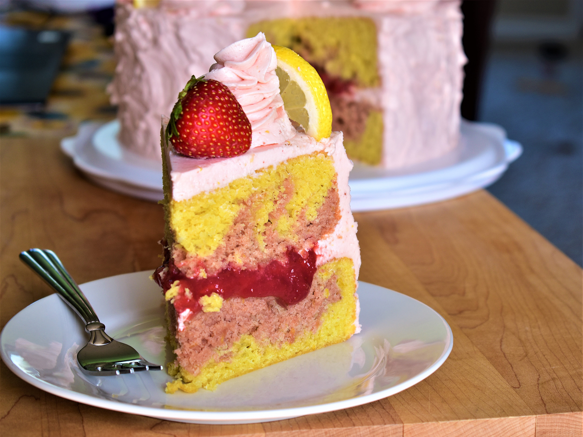 close up view of a slice of Strawberry Lemonade Cake garnished with pink cream, a fresh strawberry and a lemon slice, with Strawberry Lemonade Cake in the background