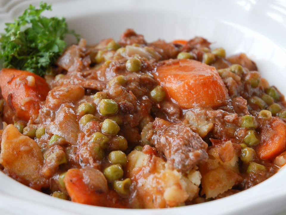 close up view of Beef Stew with beef, carrots, peas and potatoes, garnished with fresh herbs, in a white bowl