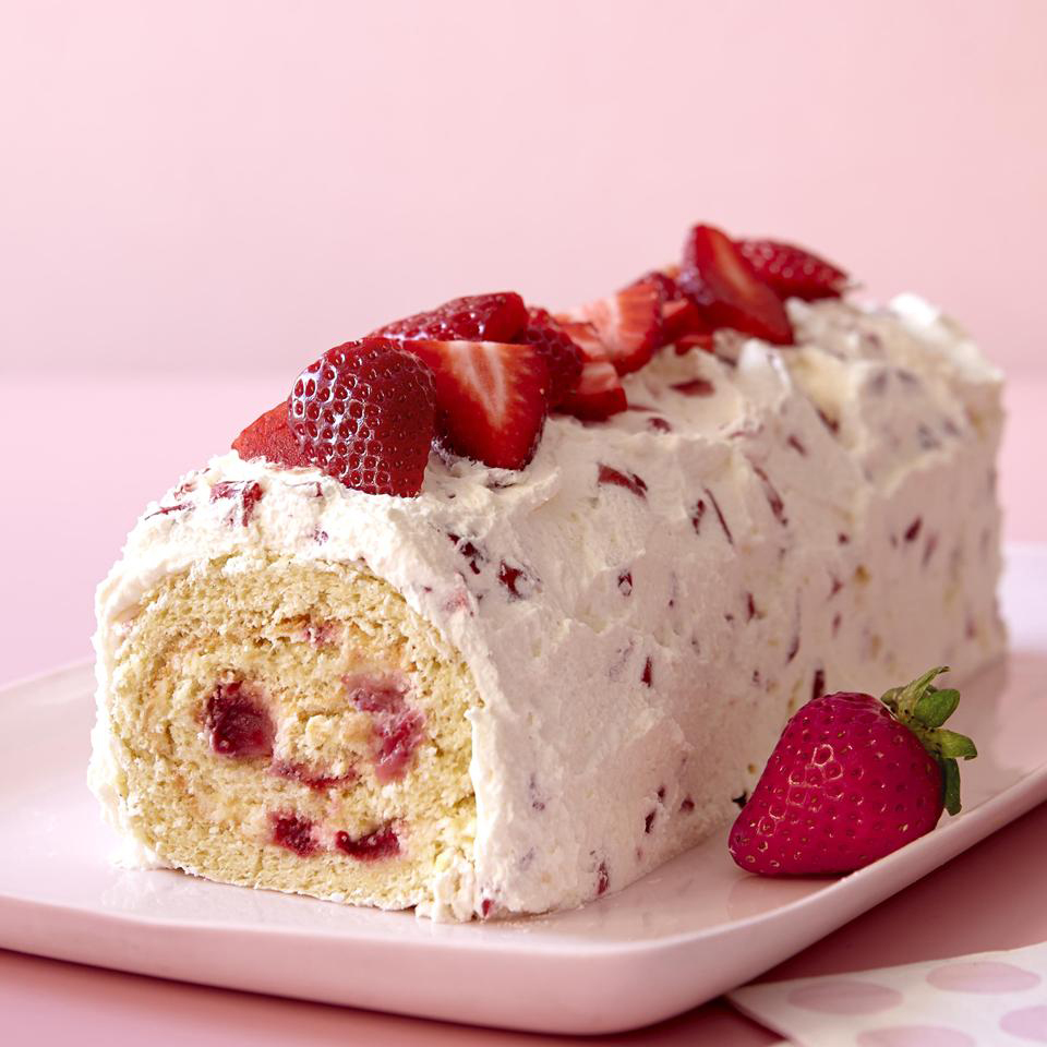close up view of a Strawberry Cream Roll, covered with strawberry cream and sliced strawberries, on a white platter with a whole strawberry, with a pink background