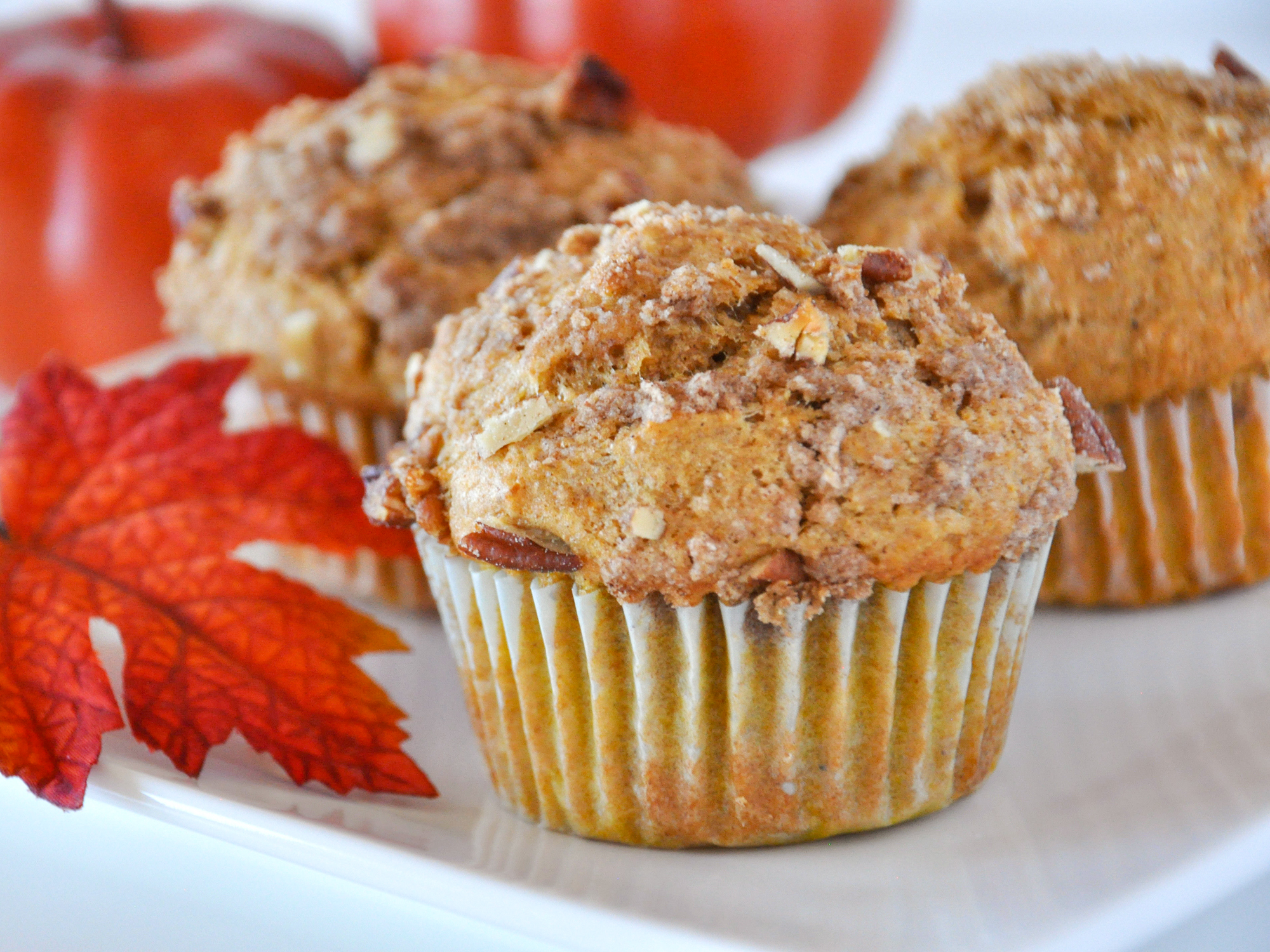 close up view of Whole Wheat Pumpkin Applesauce Muffins with nuts on top, in muffin liners, on a white plate with an orange leaf and pumpkins in the background