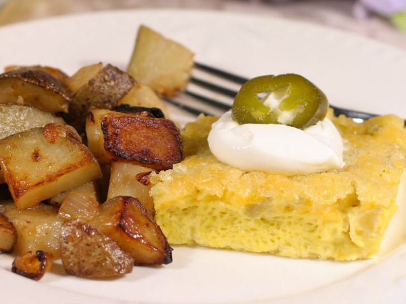 close up view of a piece of Green Chile Frittata garnished sour cream and a piece of green chile, served with roasted potatoes on a white plate with a fork
