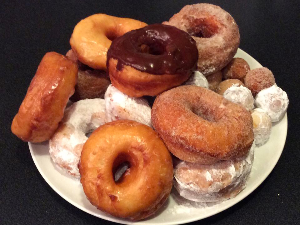 close up view of a pile of Spudnuts, donuts of various flavors including powdered sugar donuts, and chocolate covered donuts, on a white platter