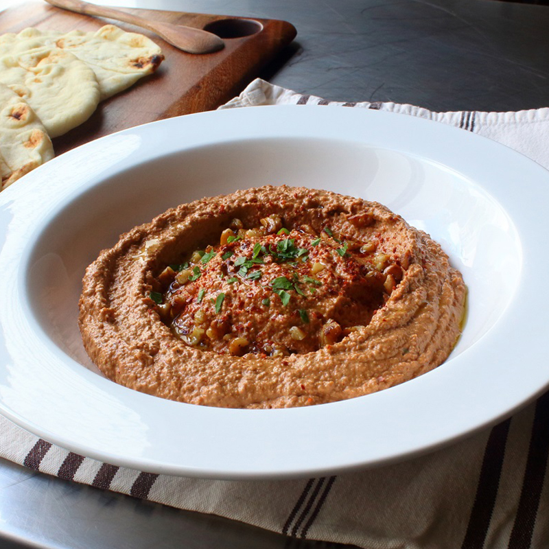 close up view of Muhammara (Roasted Pepper & Walnut Spread) garnished with fresh herbs, nuts and paprika, in a white bowl, served with pita chips