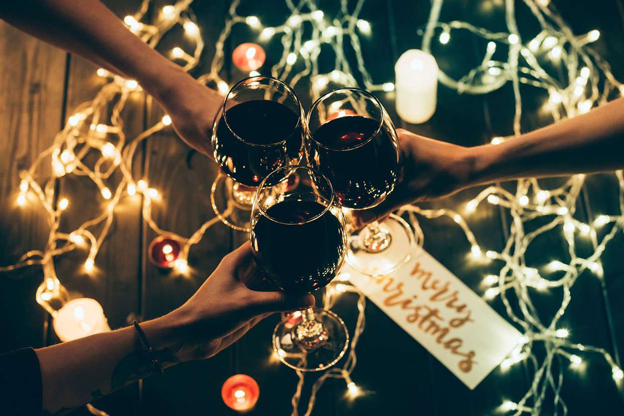 3 hands holding wine glasses together to cheers with Christmas lights and decor in the background