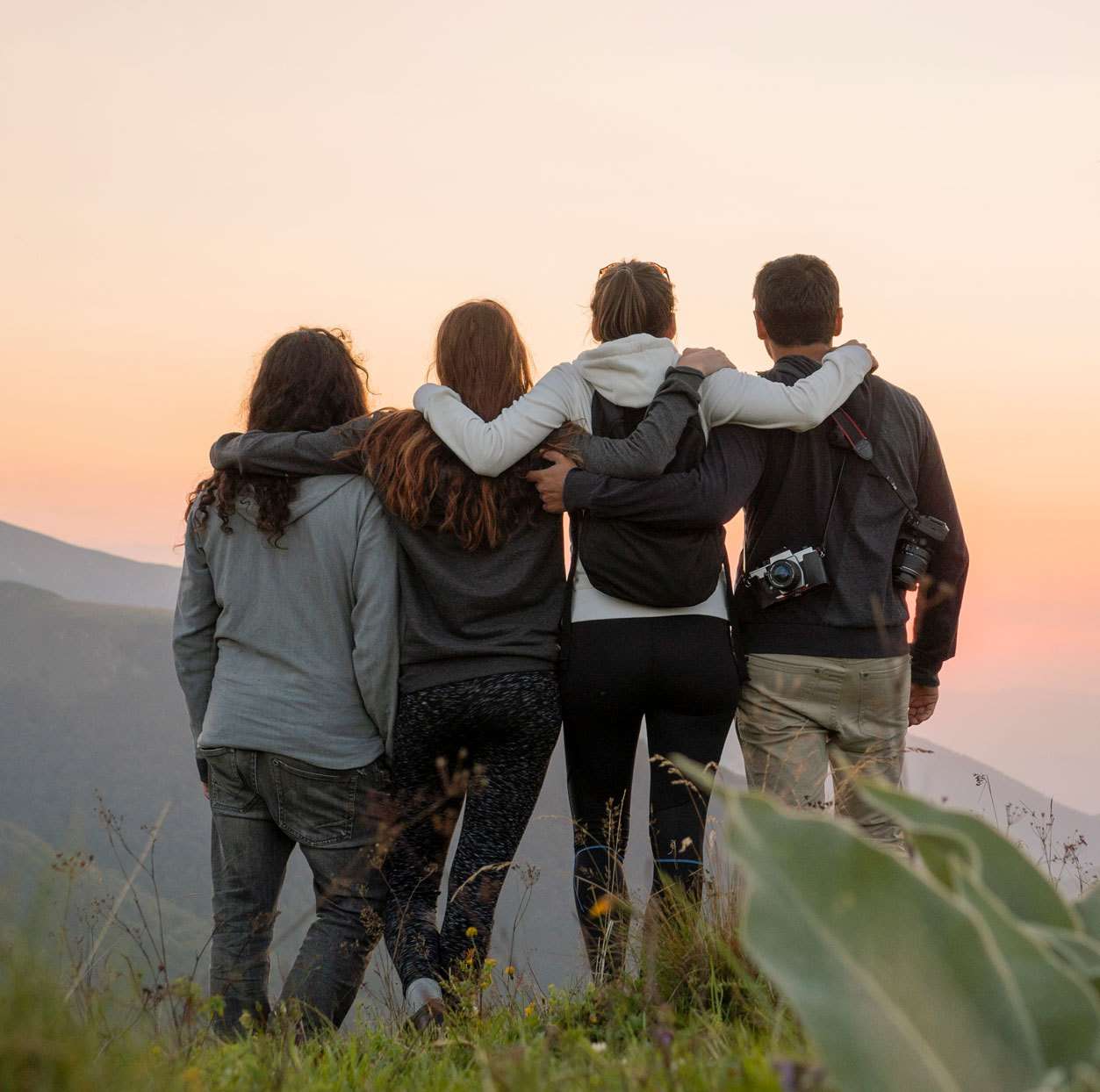 4 friends with arms around each other as they look at a sunset