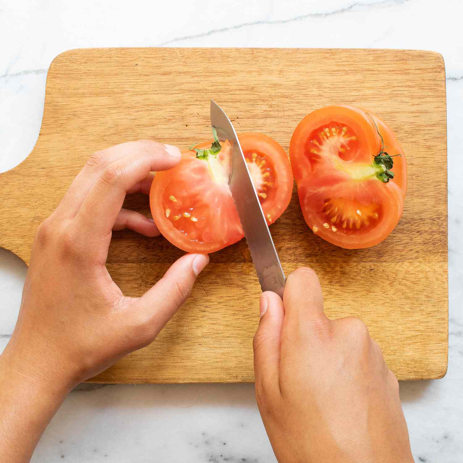 hands using a knife to cut a tomato into wedges on a wood cutting board