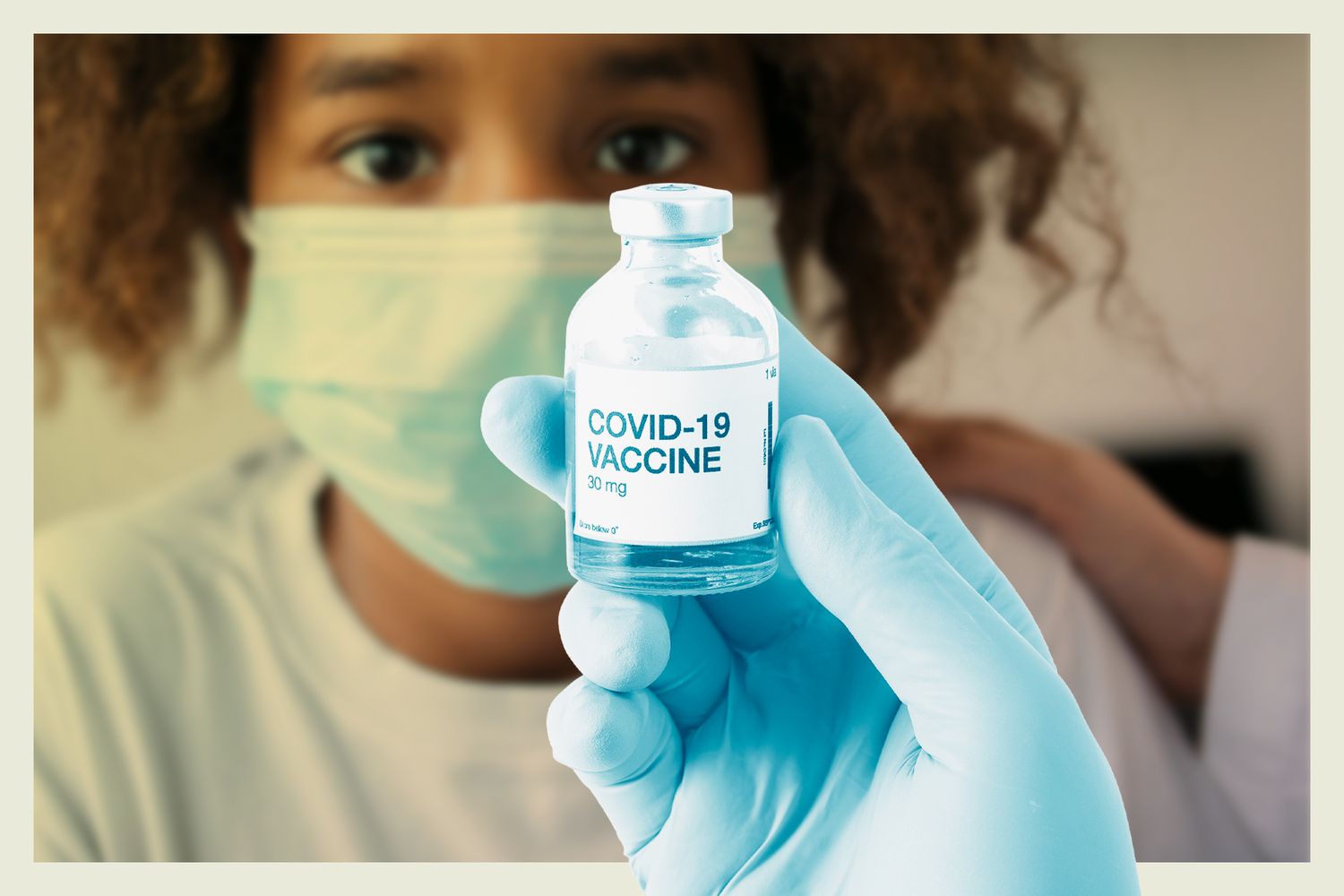 A hand holding a COVID-19 vaccine bottle with a young girl in the background wearing a mask