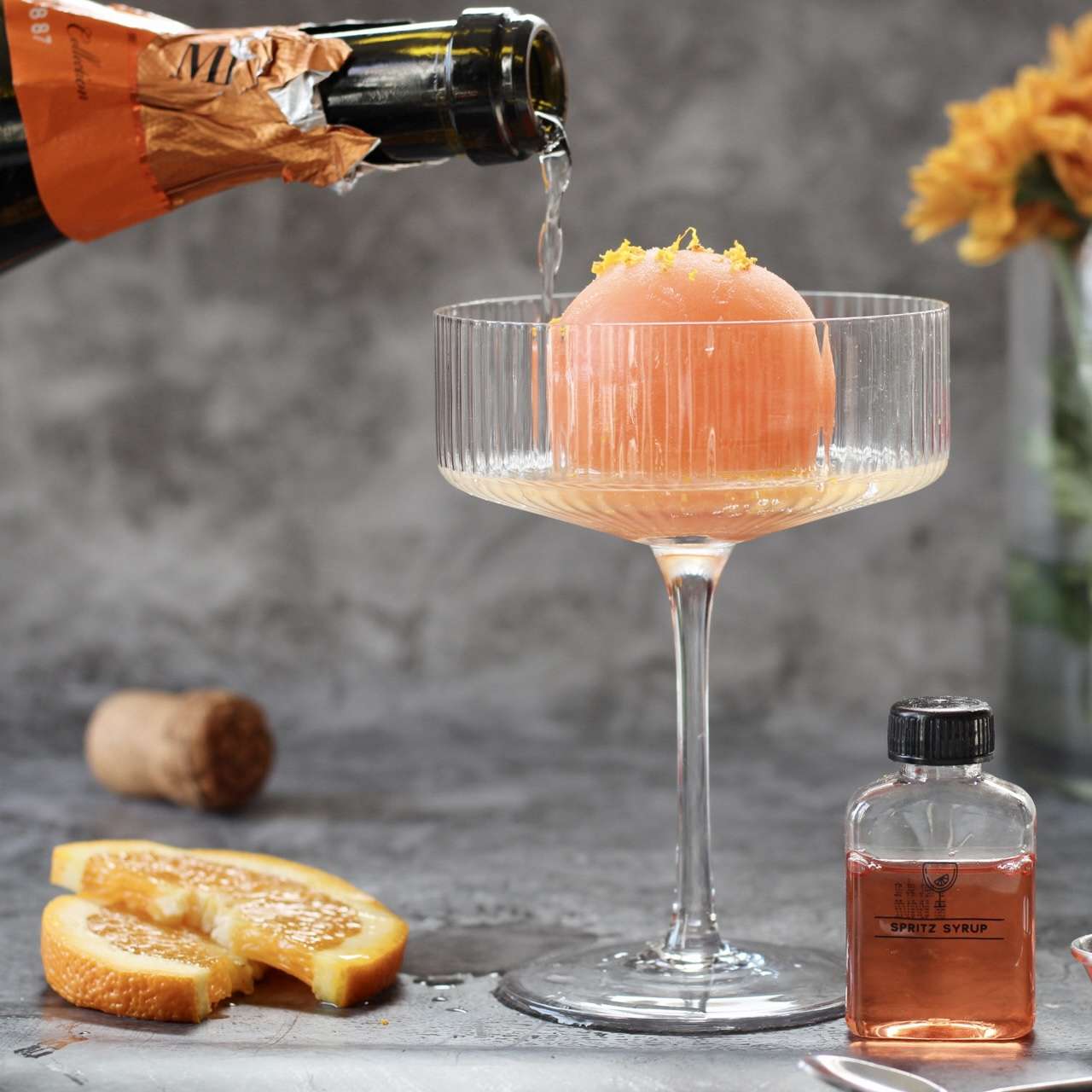 prosecco pouring into a coupe glass with a frozen pink ice cube made of juice on a grey background