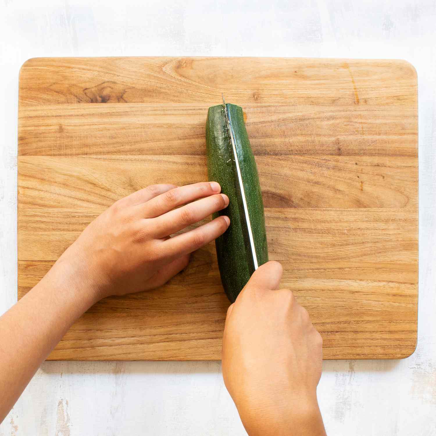 Hands using a knife to cut a zucchini on a wood cutting board