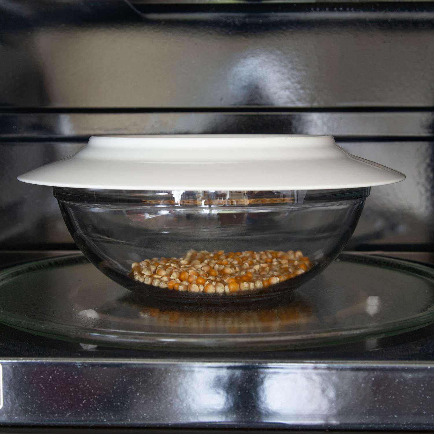 Popcorn kernels in a glass bowl with a white plate on the top in a microwave