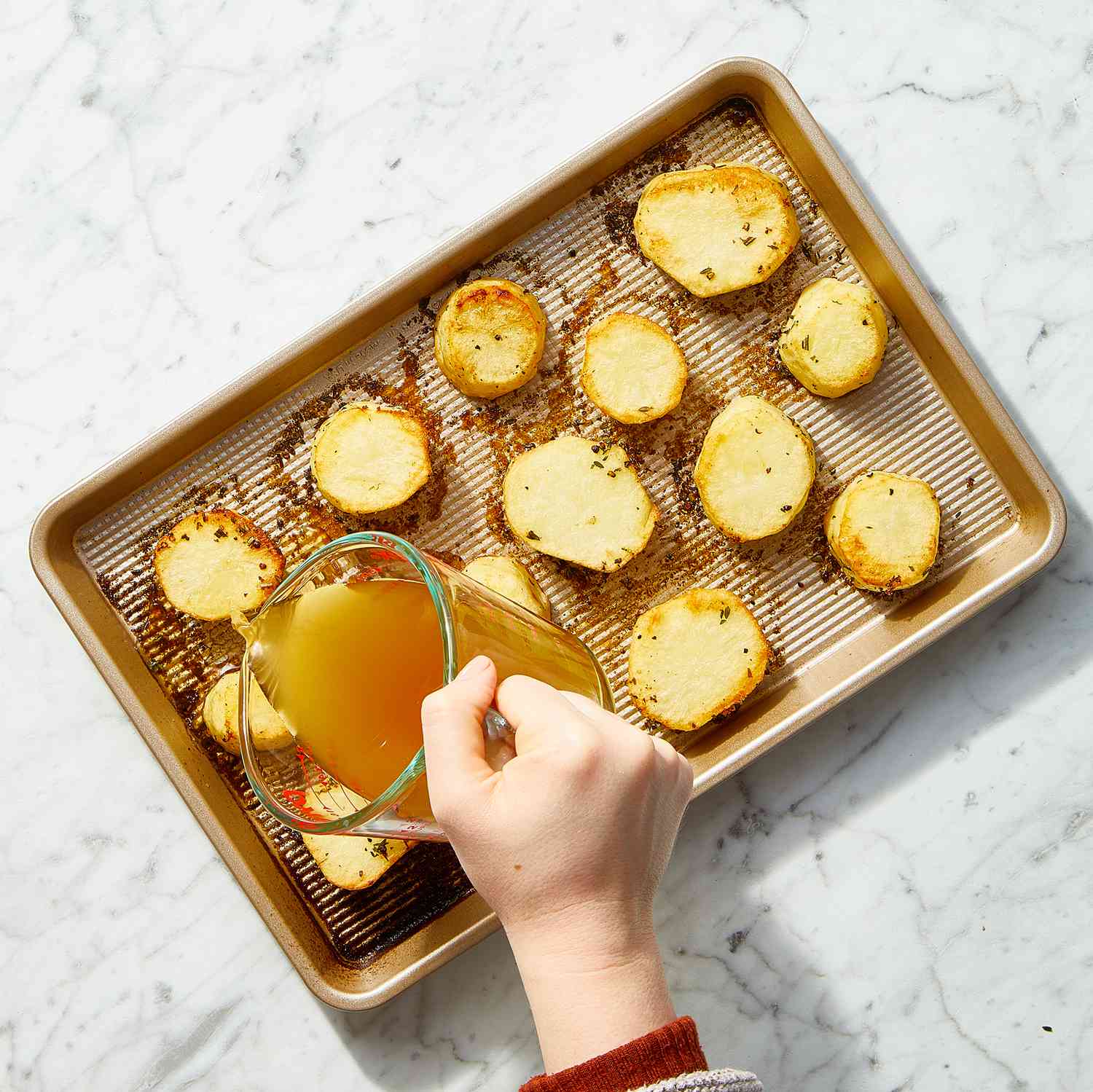 A hand pouring broth into a sheet pan with potato slices