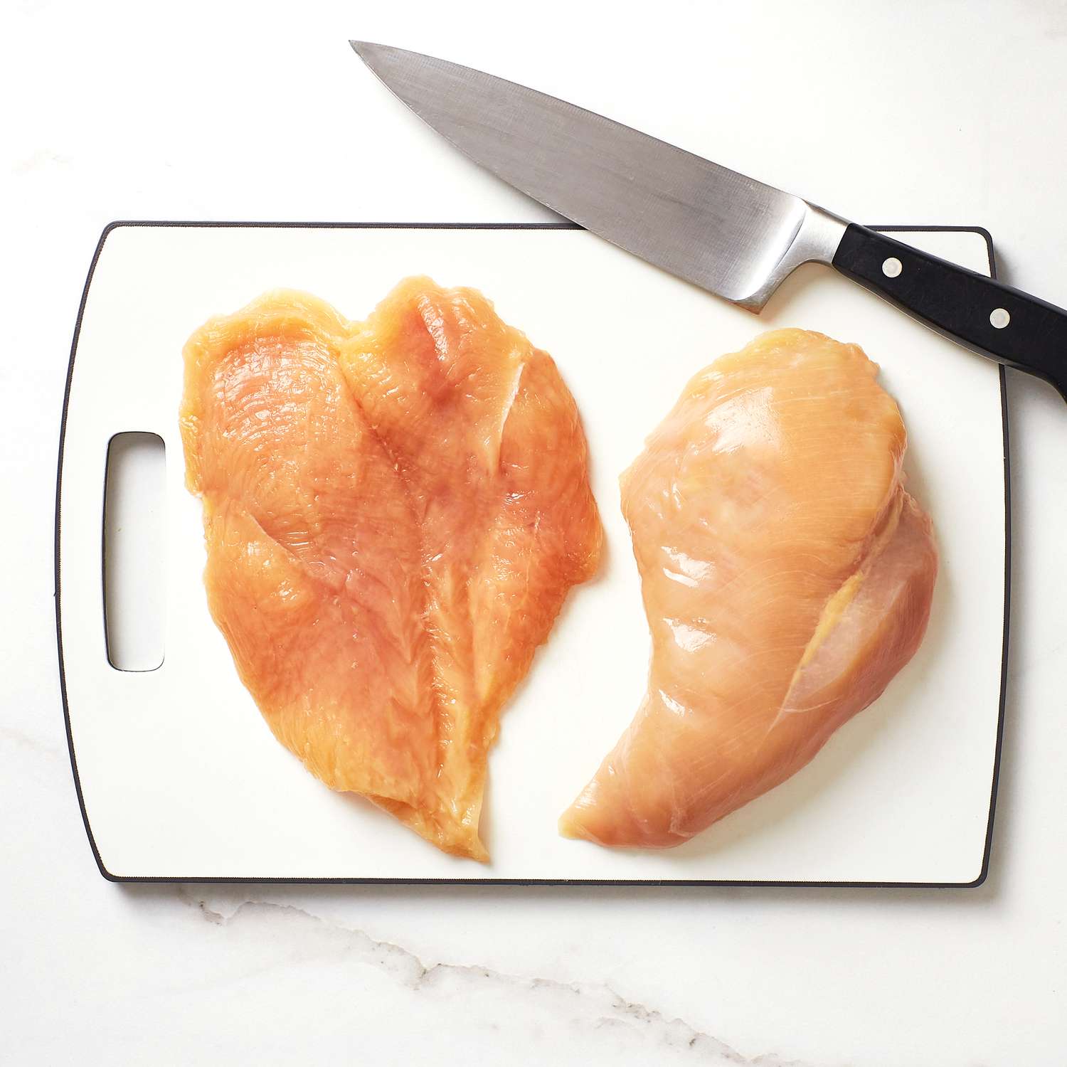 a butterflied chicken breast sitting next to a chicken breast on a white cutting board with a knife