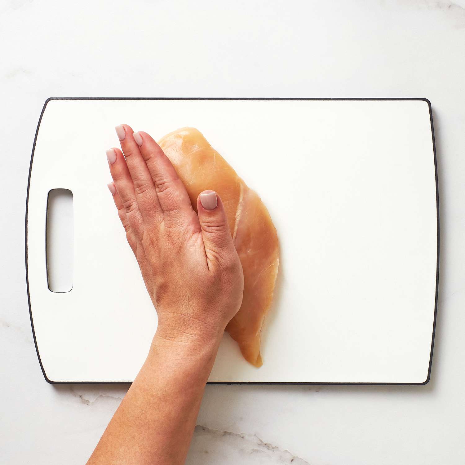 A hand placed on a chicken breast on a cutting board