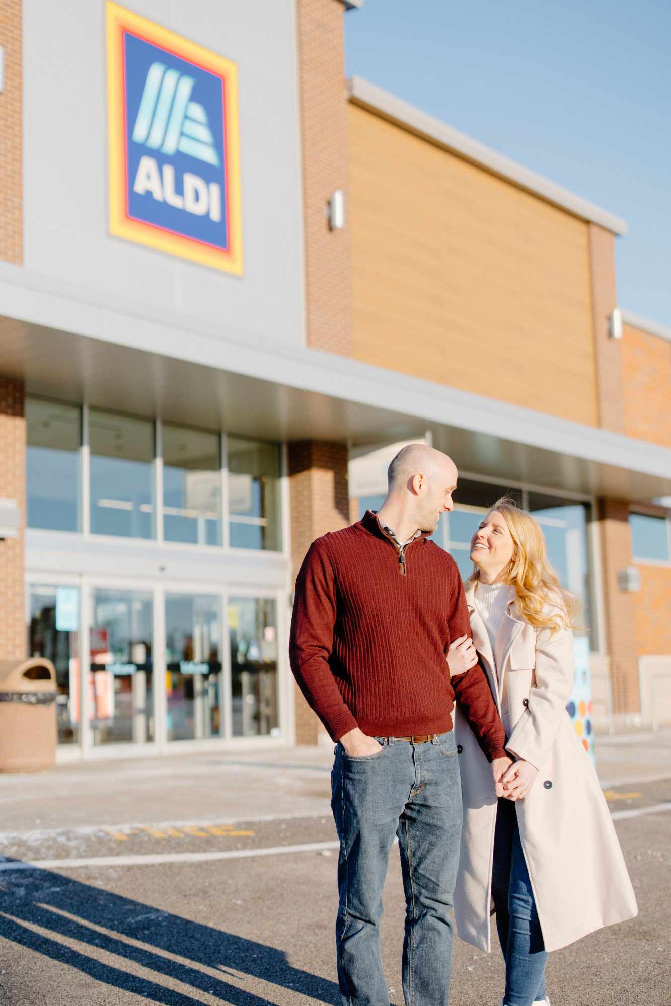 a man in a red sweater poses with a woman in a white coat in front of an Aldi store