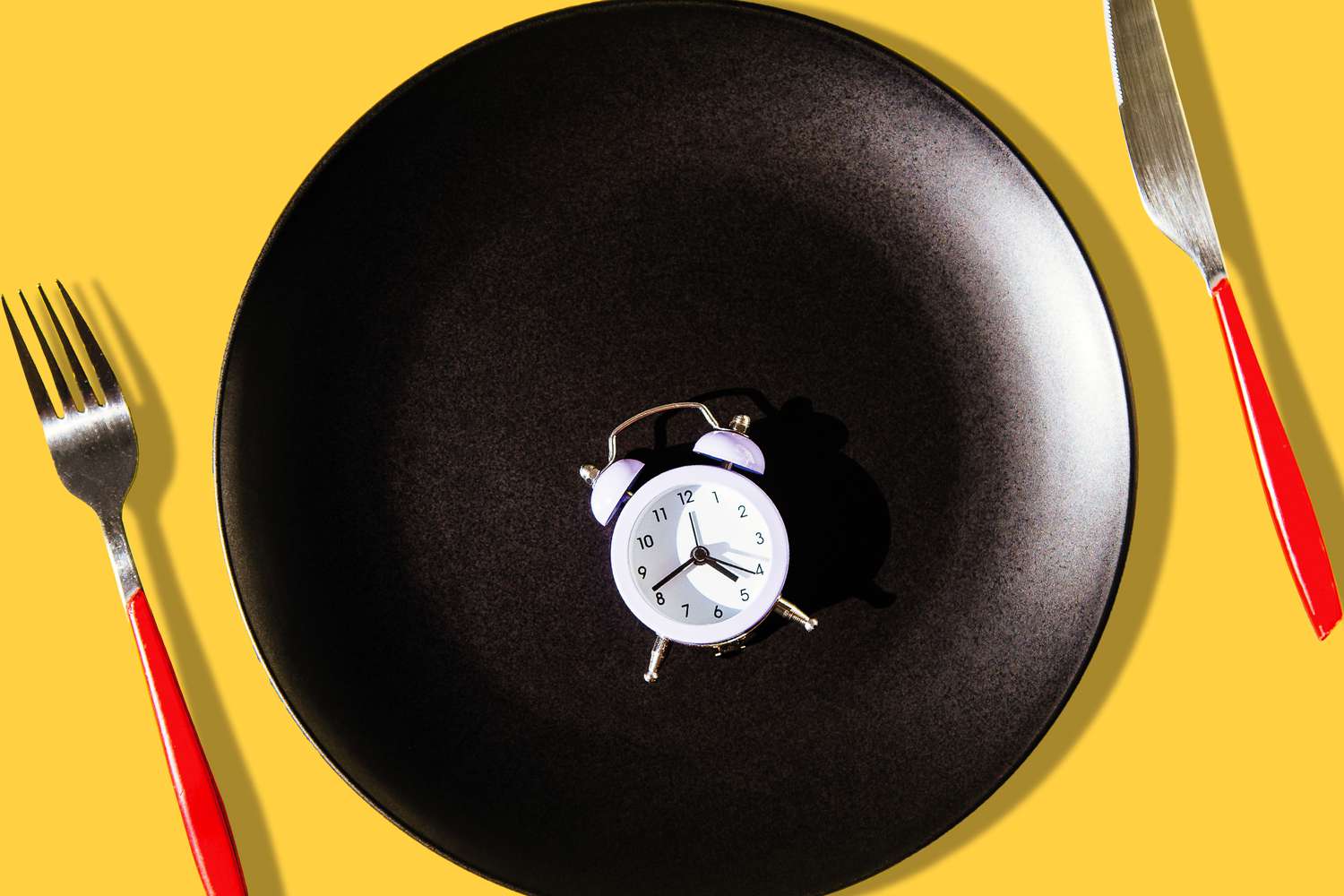 Small clock alarm on a black plate with red cutlery on yellow background