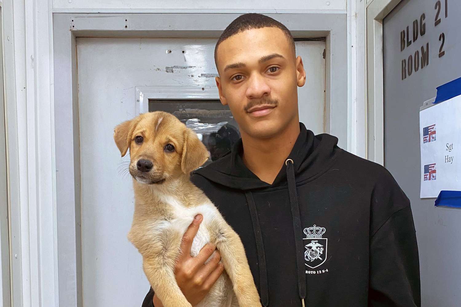 Adult black marine holds blond lab puppy in one hand